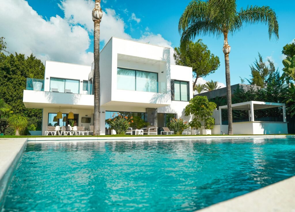 A stunning modern luxury villa with a pool, surrounded by lush greenery and bathed in sunlight.